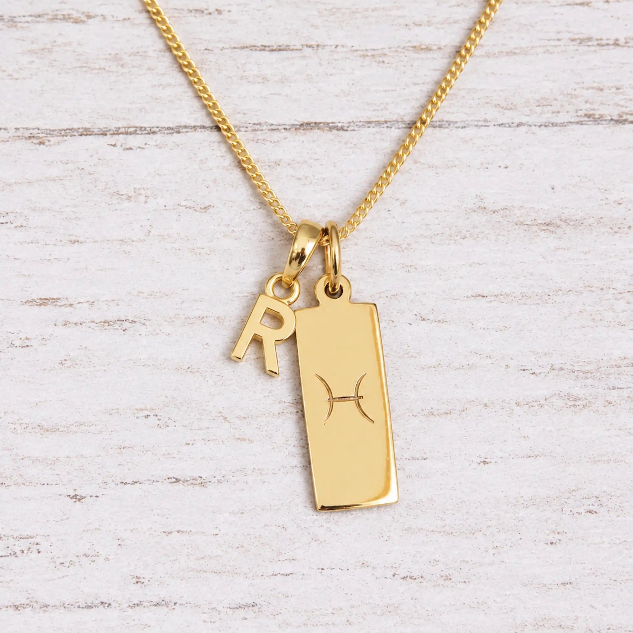 Gold Pisces initial necklace with a letter R charm and engraved zodiac bar on white wood, close-up