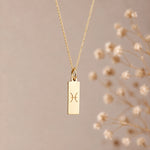 A gold pisces necklace featuring a rectangular pendant with an engraved Pisces zodiac symbol, hanging from a delicate gold chain against a soft beige background with blurred dried flowers on the right.