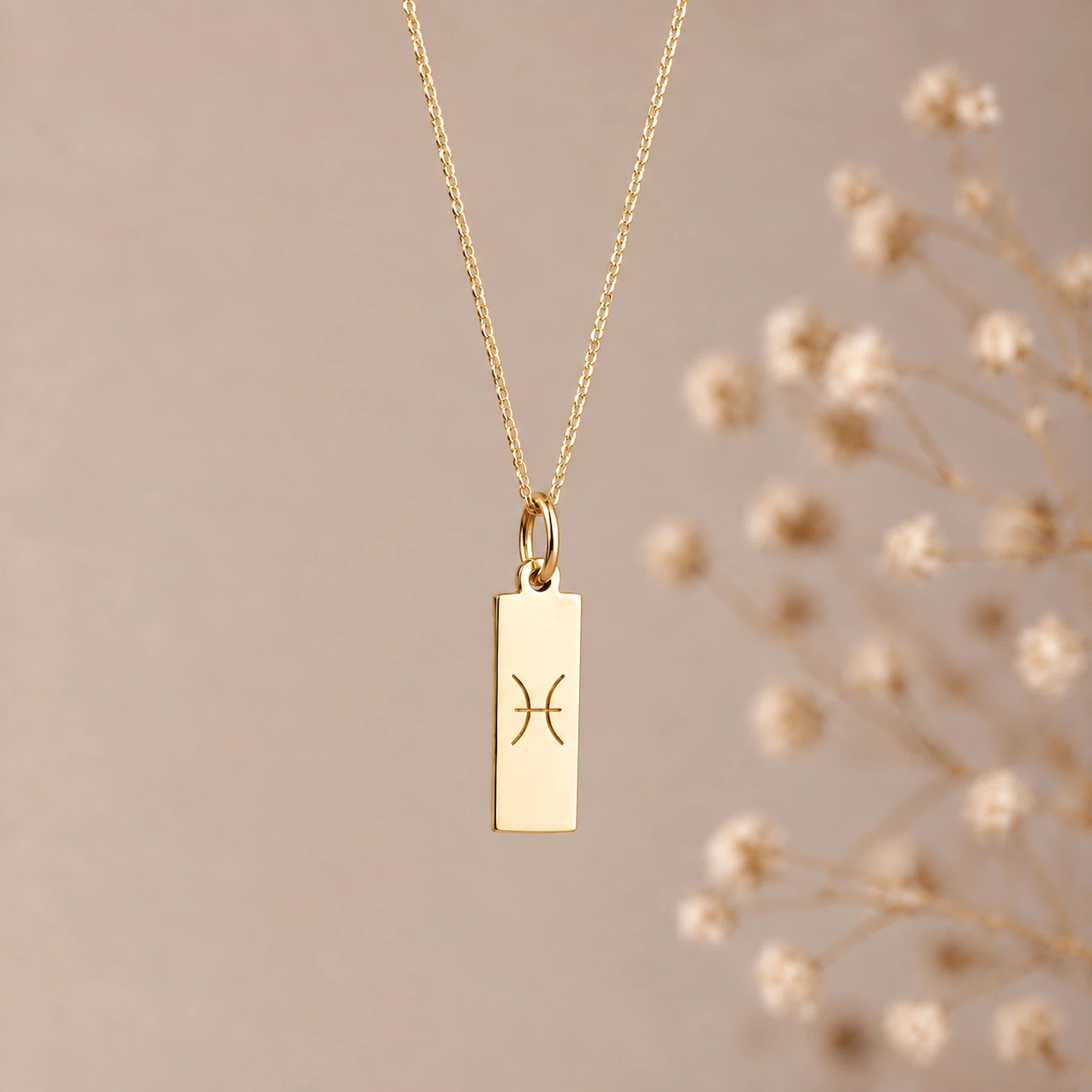 A gold pisces necklace featuring a rectangular pendant with an engraved Pisces zodiac symbol, hanging from a delicate gold chain against a soft beige background with blurred dried flowers on the right.