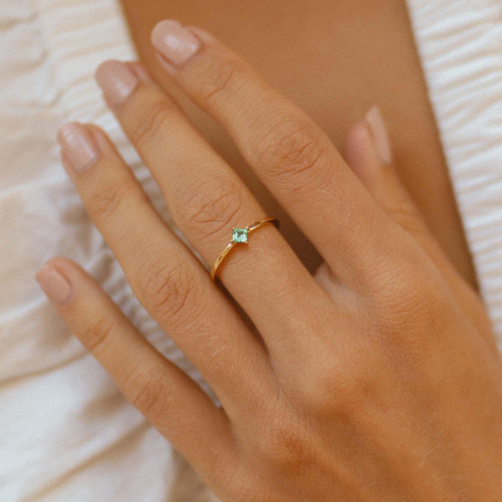Hand wearing a gold ring with a green gemstone on a neutral background
