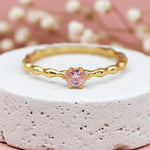 Gold ring with a pink rose quartz gemstone on a textured surface with blurred flowers in the background