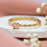 Gold ring with a pink rose quartz gemstone on a white stone surface with blurred flowers in the background