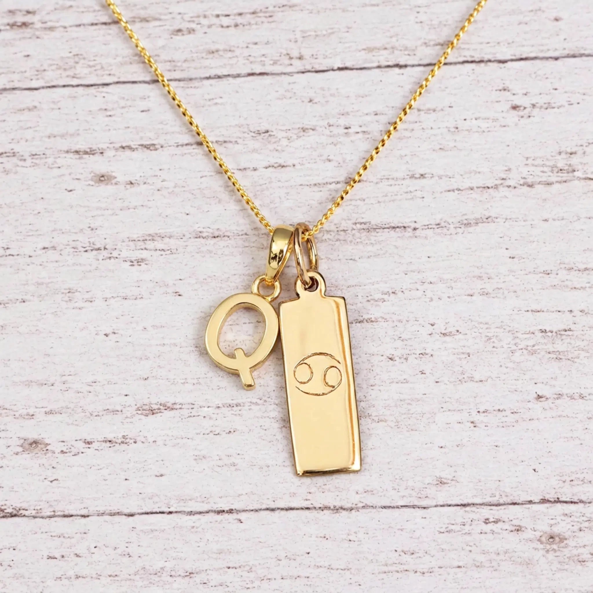 Gold Initial Q Pendant and Cancer Zodiac Pendant Necklace on a fine gold chain against a light wooden background