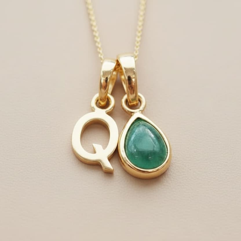 Gold ‘Q’ initial necklace with a green emerald teardrop pendant on a beige background