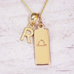 Gold initial and Libra zodiac necklace with a letter R charm and engraved bar pendant on whitewashed wood