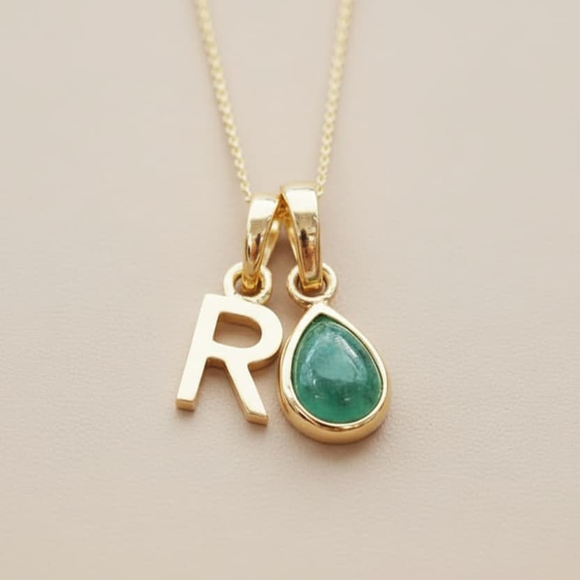 Gold necklace with 'R' pendant and green natural emerald teardrop pendant on a beige background