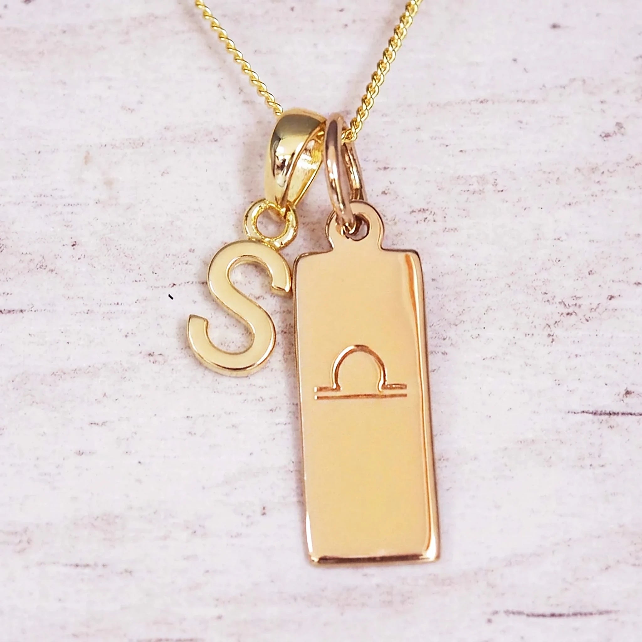 Gold initial and Libra zodiac necklace with a letter S charm and engraved bar pendant on whitewashed wood
