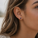A close-up of a woman's ear wearing a september birthstone natural sapphire earring - small teardrop-shaped stud earring with a deep blue stone in a gold-toned setting.