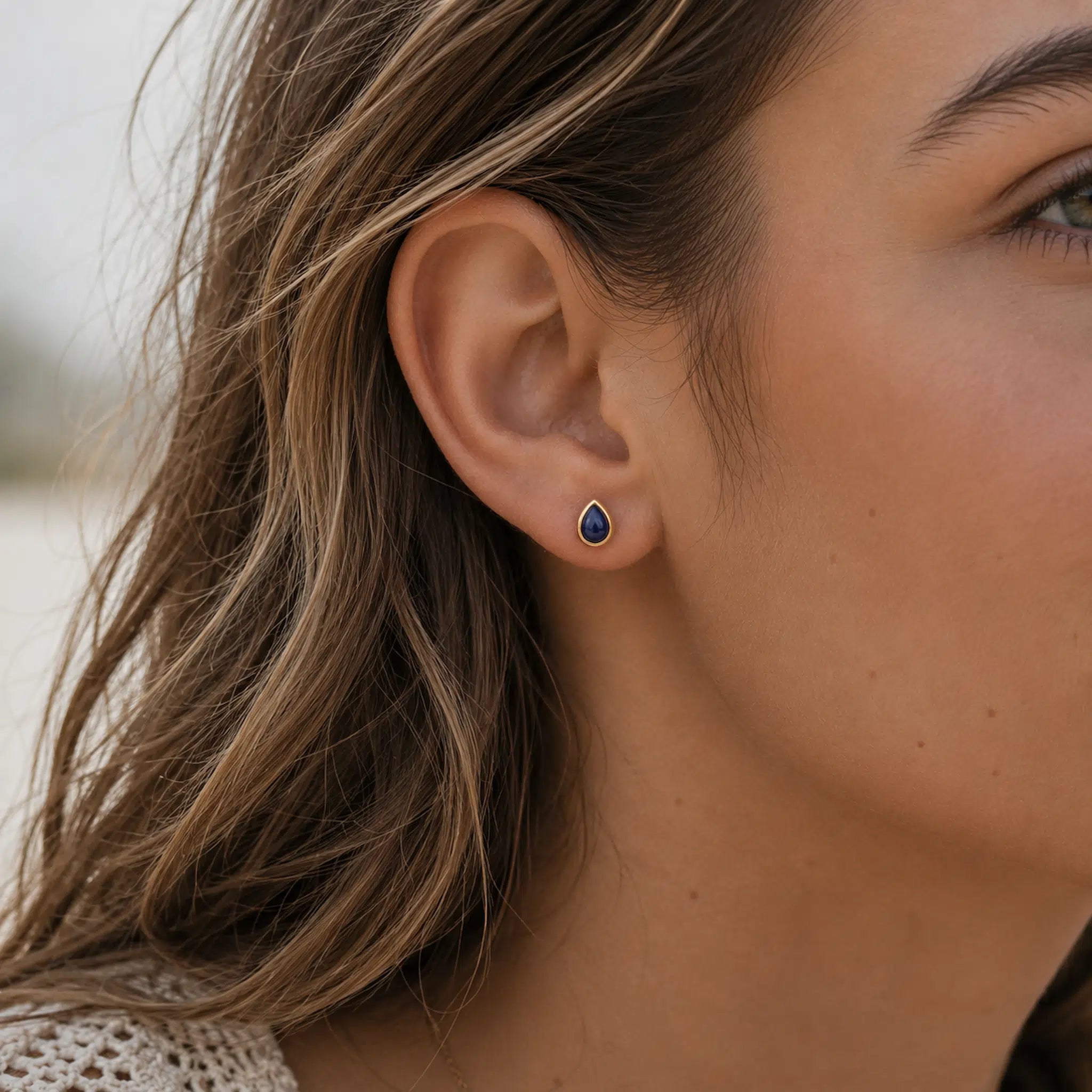 A close-up of a woman's ear wearing a september birthstone natural sapphire earring - small teardrop-shaped stud earring with a deep blue stone in a gold-toned setting.