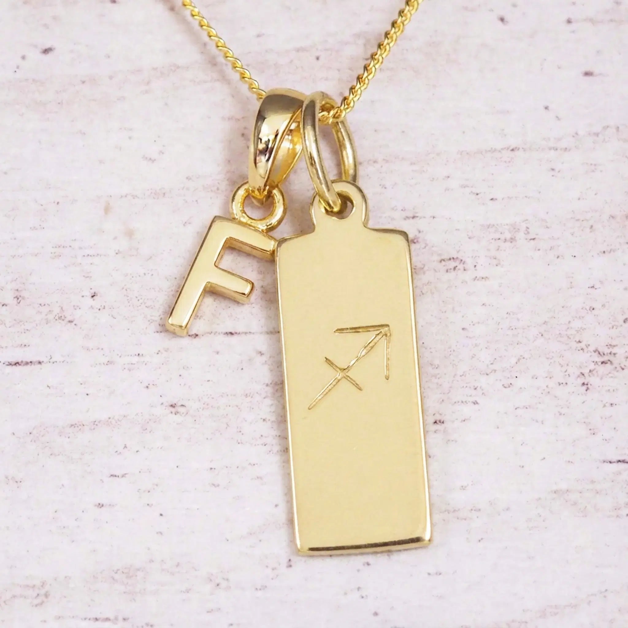 Gold Sagittarius initial necklace with a letter F charm and engraved zodiac bar on white wood, close-up