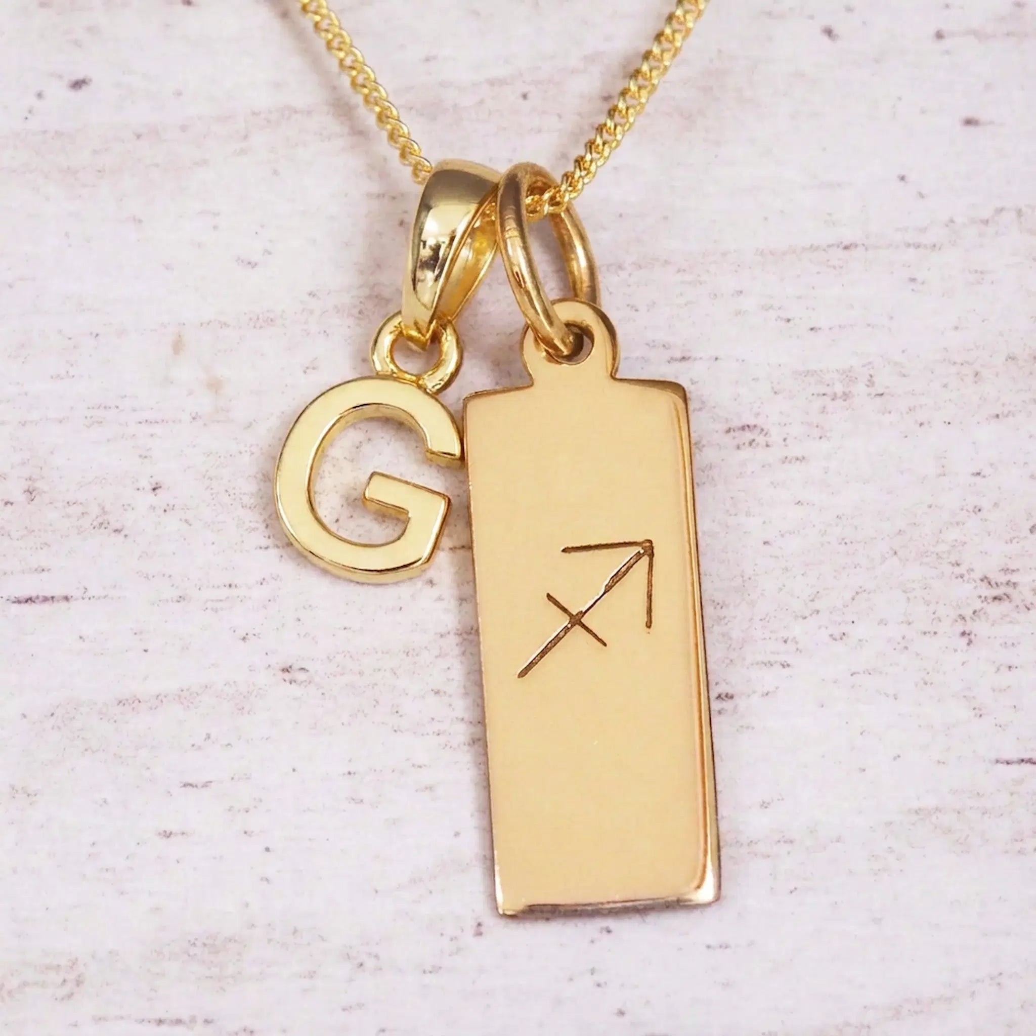 Gold Sagittarius initial necklace with a letter G charm and engraved zodiac bar on white wood, close-up