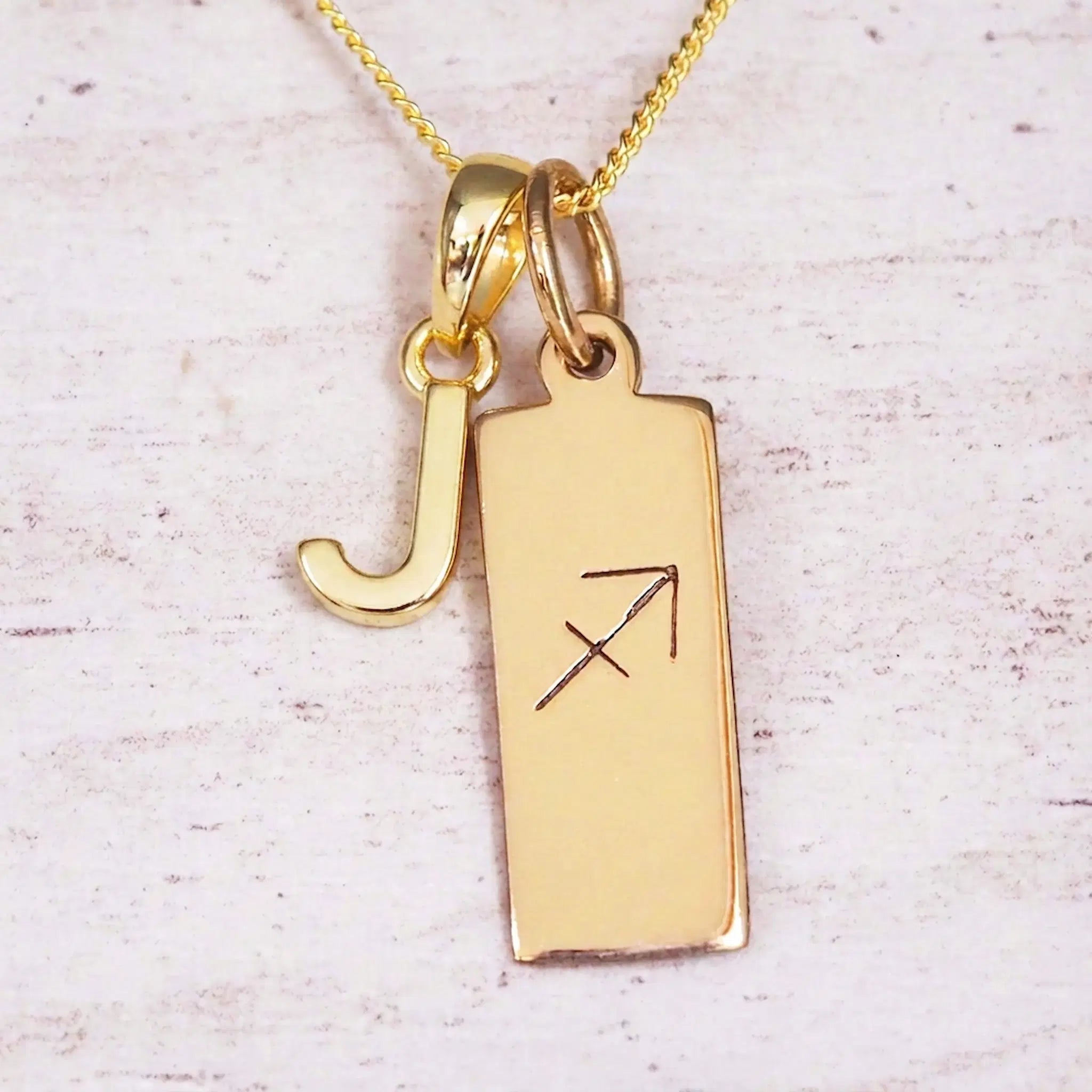 Gold Sagittarius initial necklace with a letter J charm and engraved zodiac bar on white wood, close-up