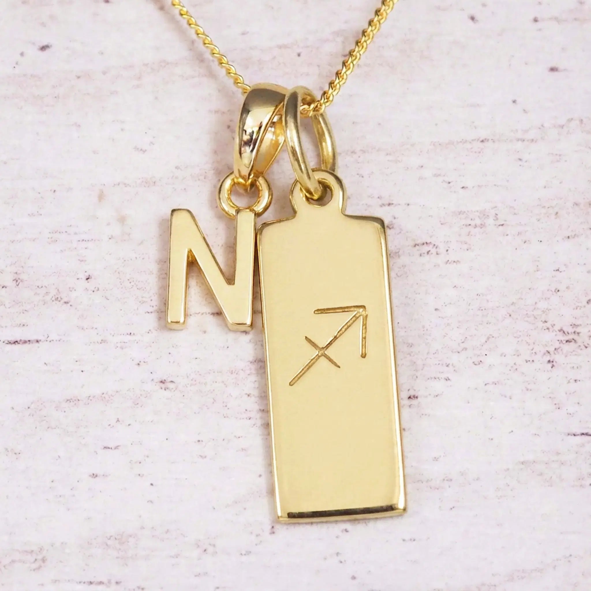 Gold Sagittarius initial necklace with a letter N charm and engraved zodiac bar on white wood, close-up