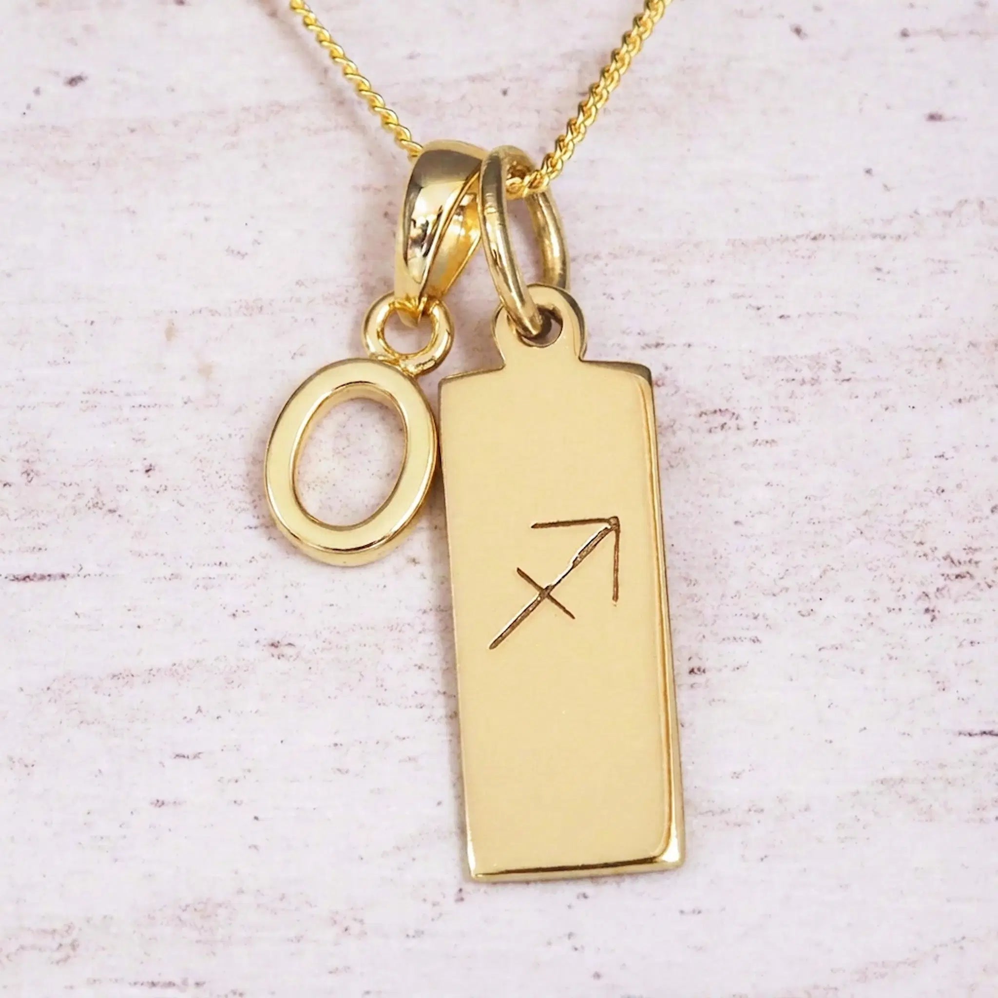 Gold Sagittarius initial necklace with a letter O charm and engraved zodiac bar on white wood, close-up