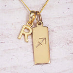 Gold Sagittarius initial necklace with a letter R charm and engraved zodiac bar on white wood, close-up