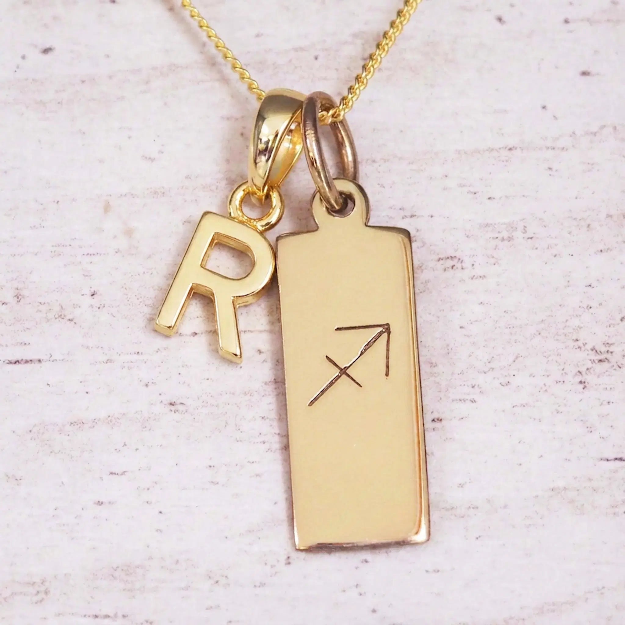 Gold Sagittarius initial necklace with a letter R charm and engraved zodiac bar on white wood, close-up