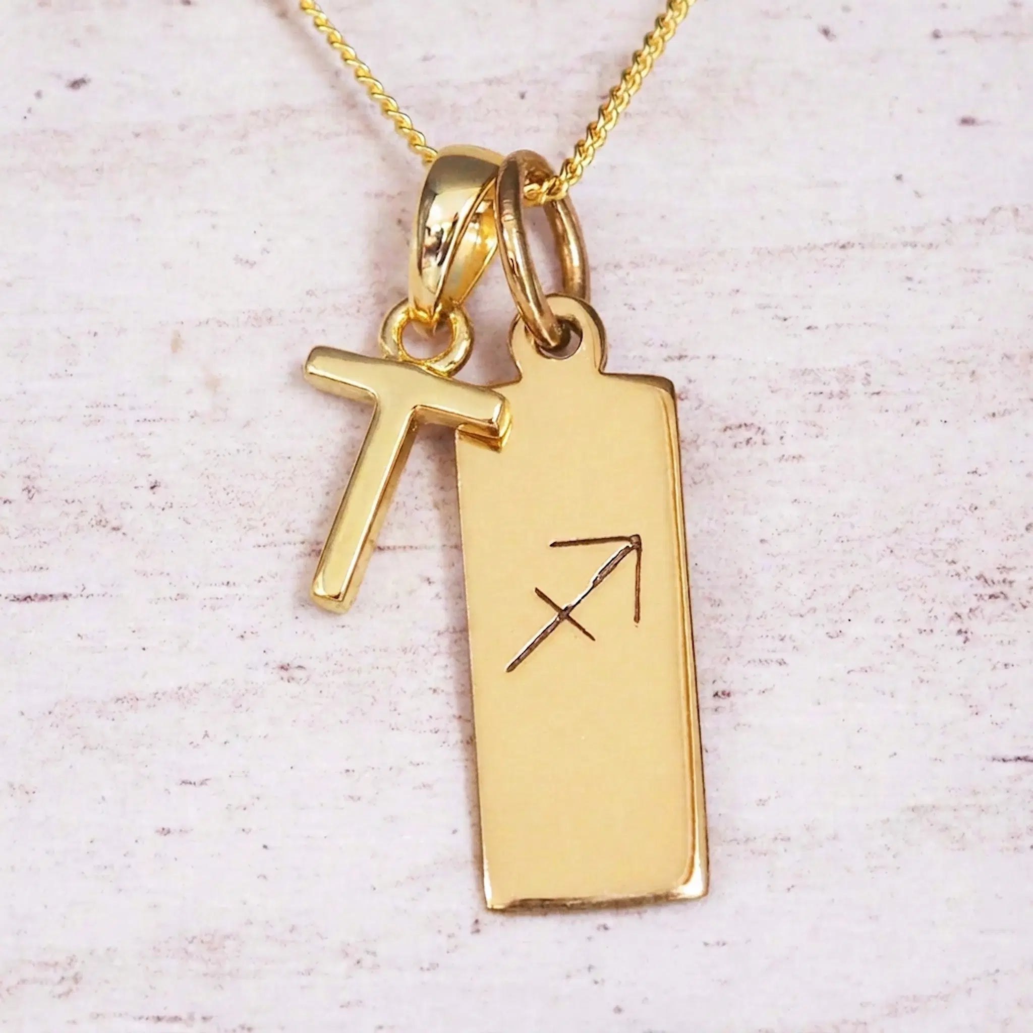 Gold Sagittarius initial necklace with a letter T charm and engraved zodiac bar on white wood, close-up