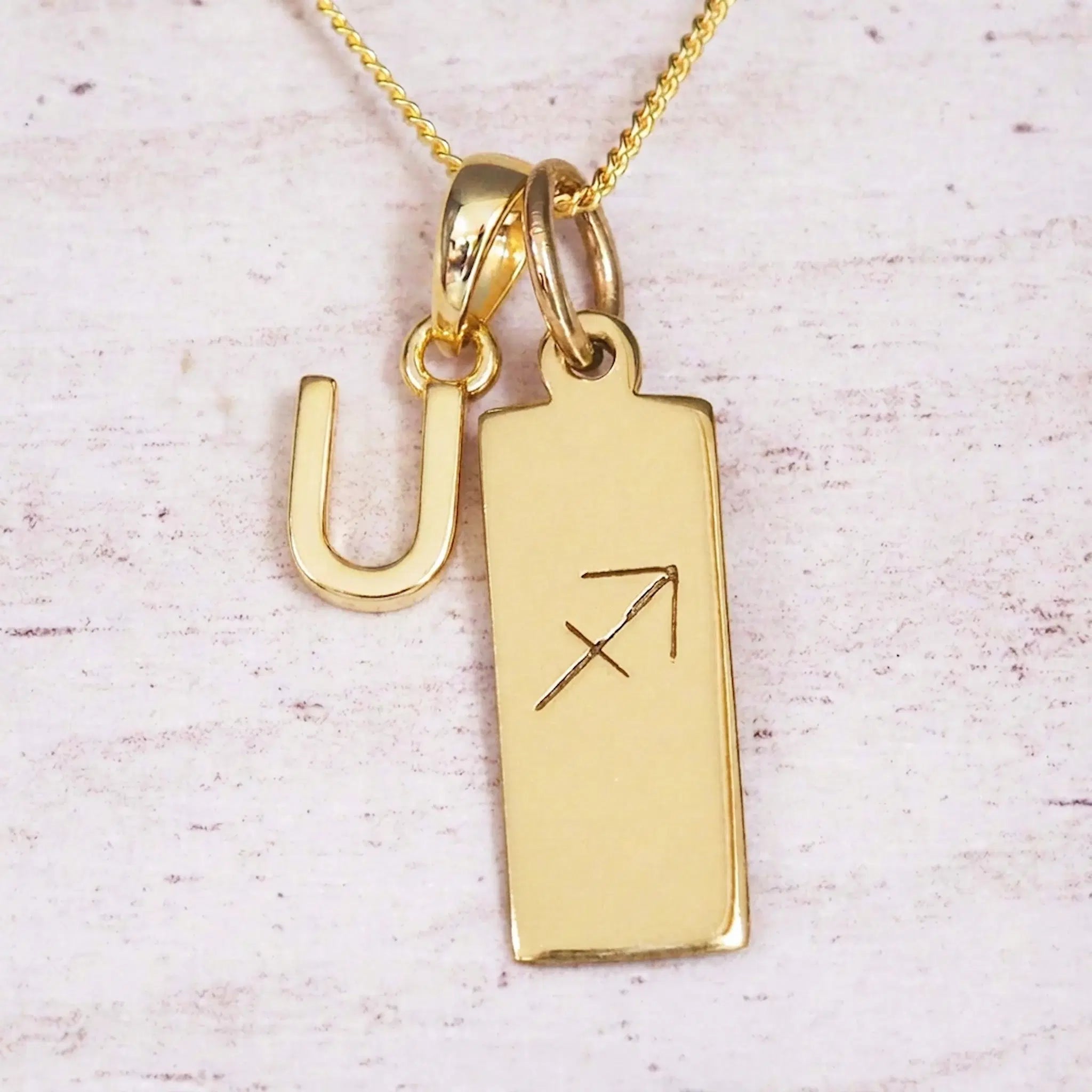 Gold Sagittarius initial necklace with a letter U charm and engraved zodiac bar on white wood, close-up