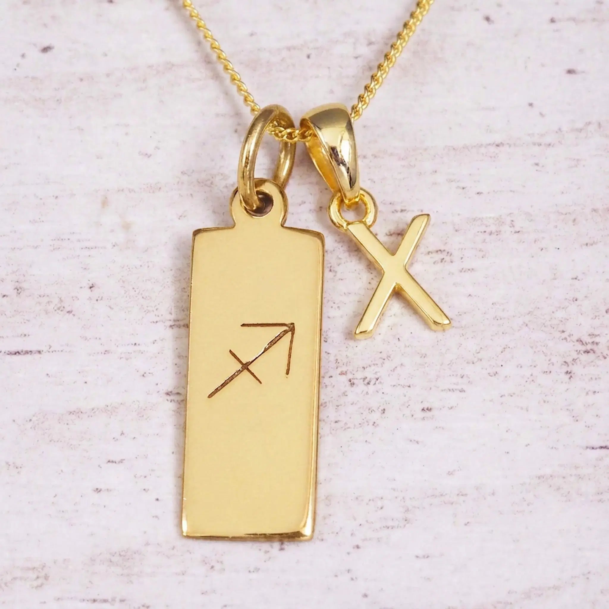 Gold Sagittarius initial necklace with a letter X charm and engraved zodiac bar on white wood, close-up