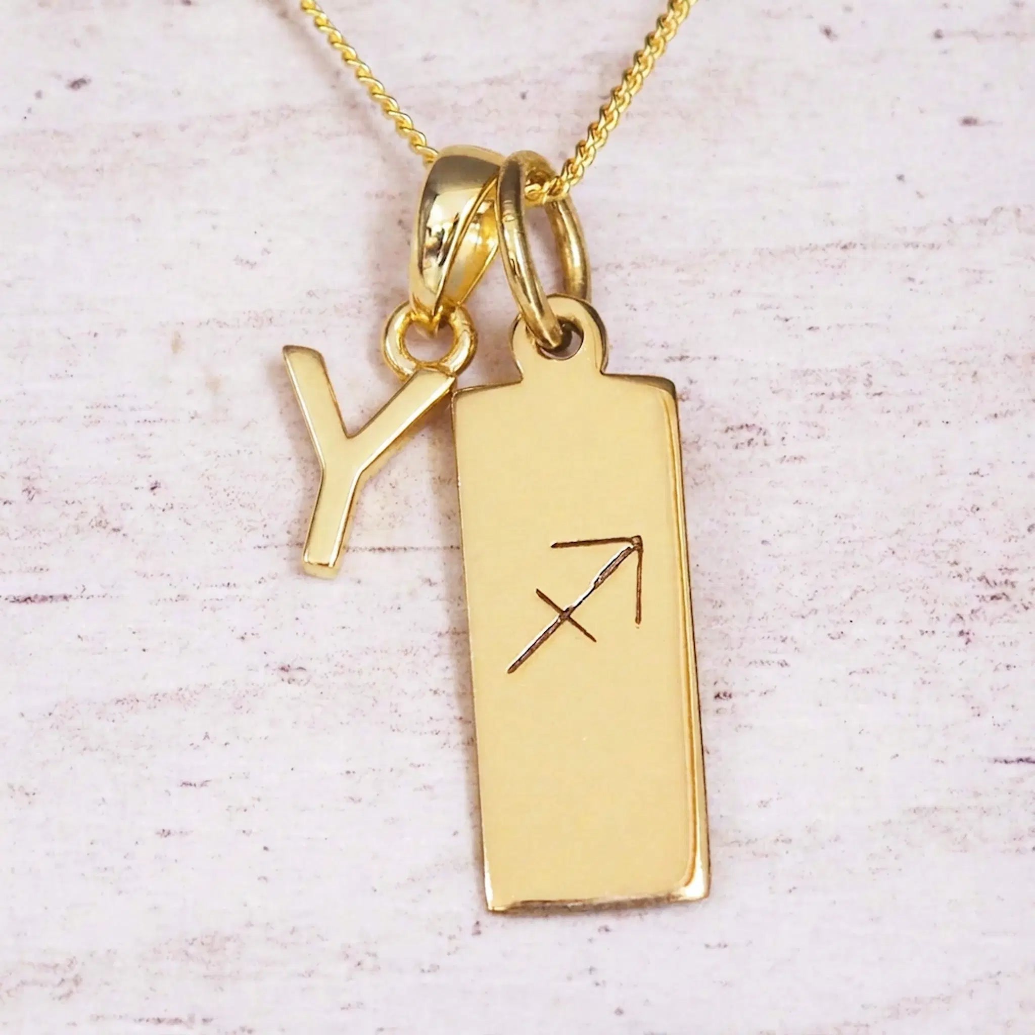 Gold Sagittarius initial necklace with a letter Y charm and engraved zodiac bar on white wood, close-up