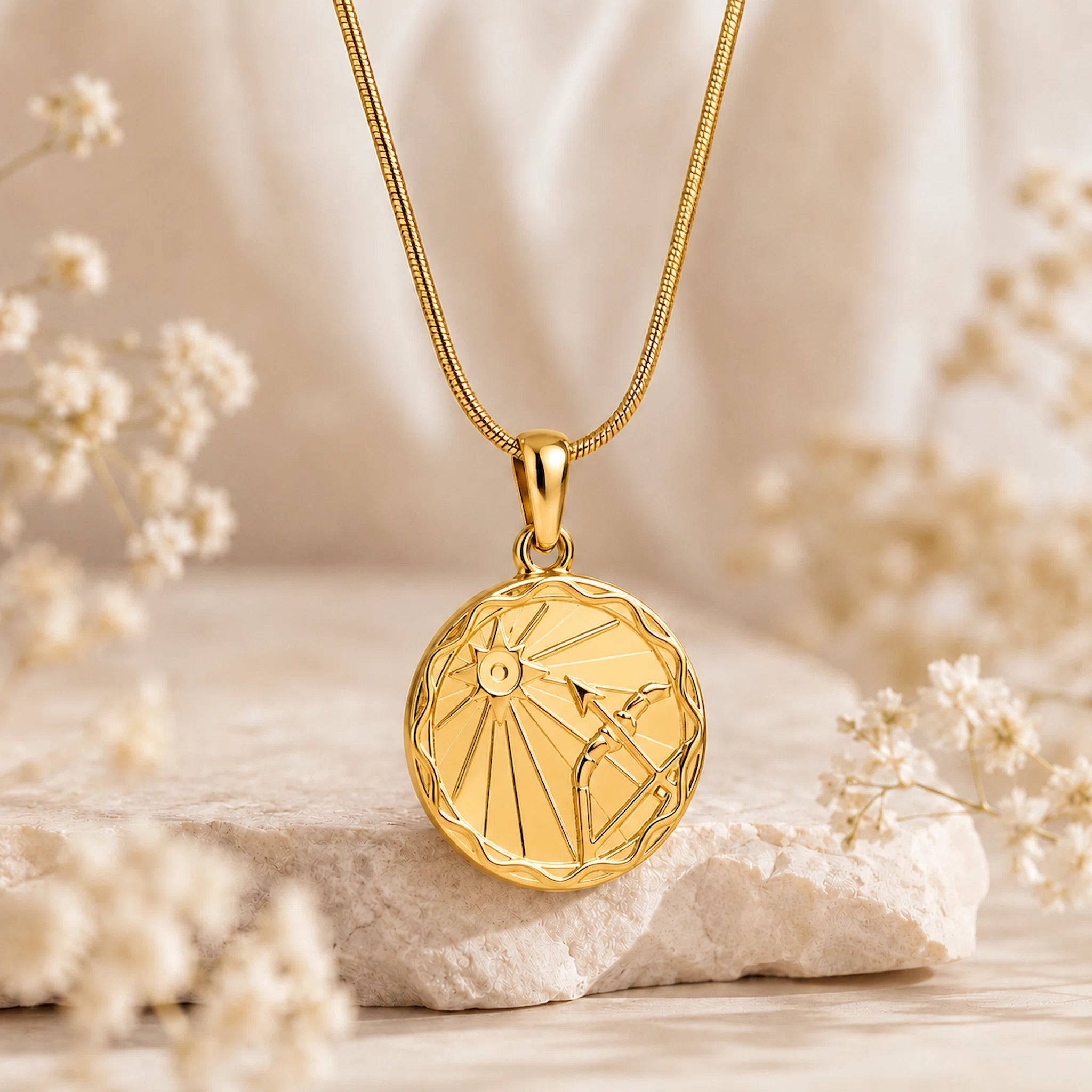 A gold pendant necklace with a circular sagittarius zodiac sign design, resting on a textured stone with soft dried flowers in a warm, neutral setting.