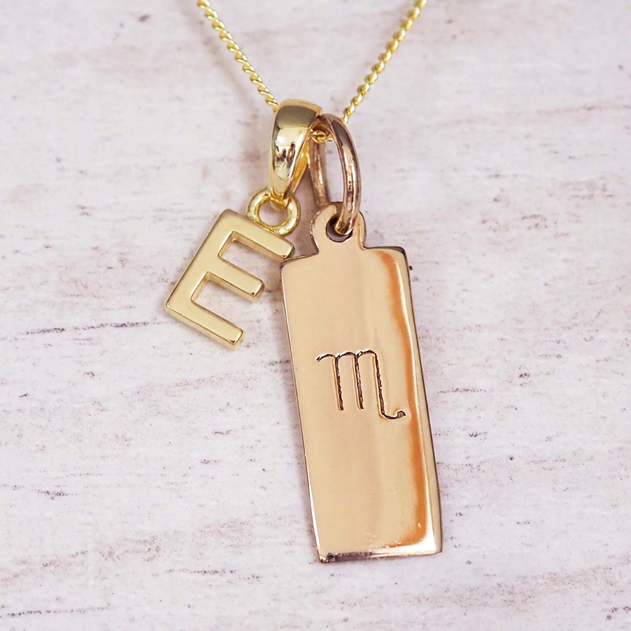 Gold Scorpio initial necklace with a letter E charm and engraved zodiac bar on white wood, close-up
