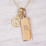 Gold Scorpio initial necklace with a letter G charm and engraved zodiac bar on white wood, close-up