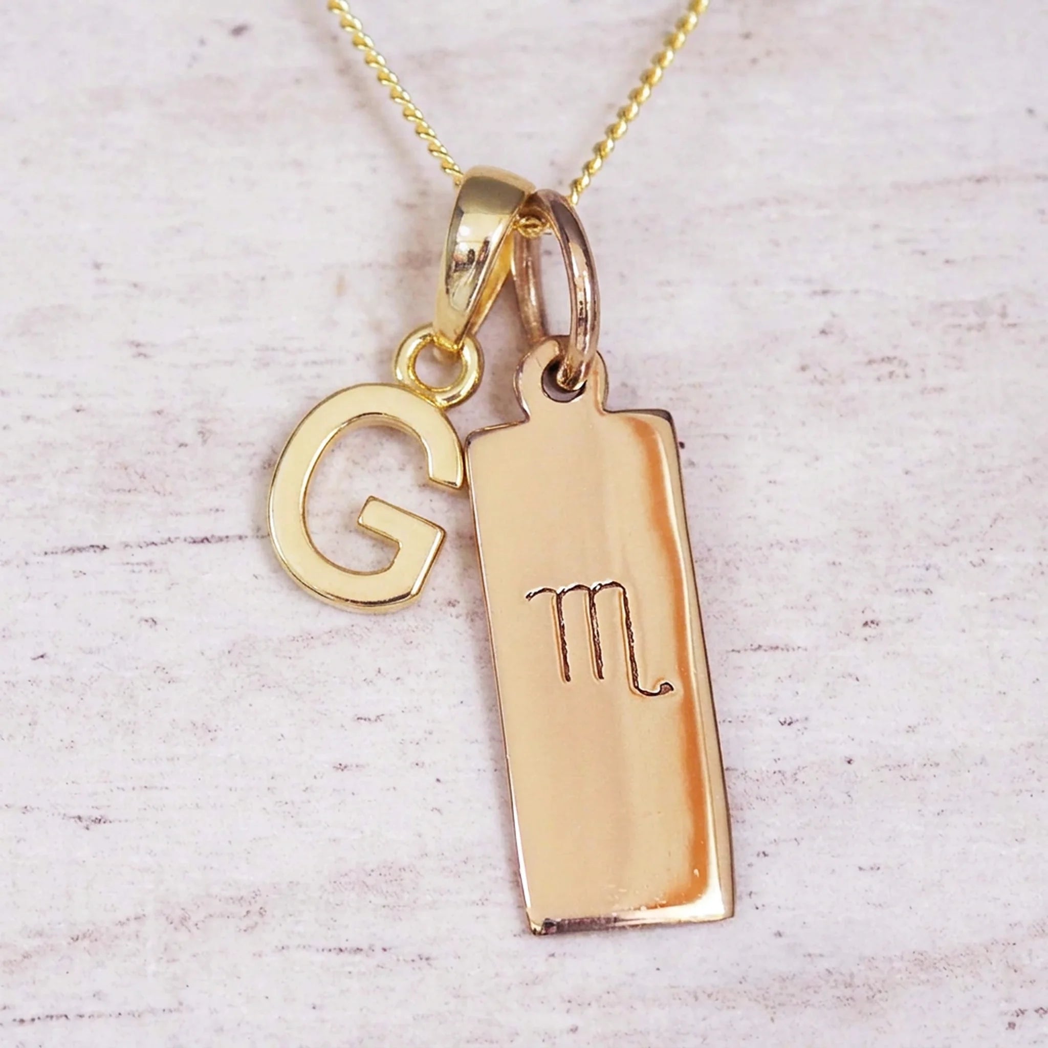 Gold Scorpio initial necklace with a letter G charm and engraved zodiac bar on white wood, close-up