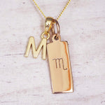 Gold Scorpio initial necklace with a letter M charm and engraved zodiac bar on white wood, close-up