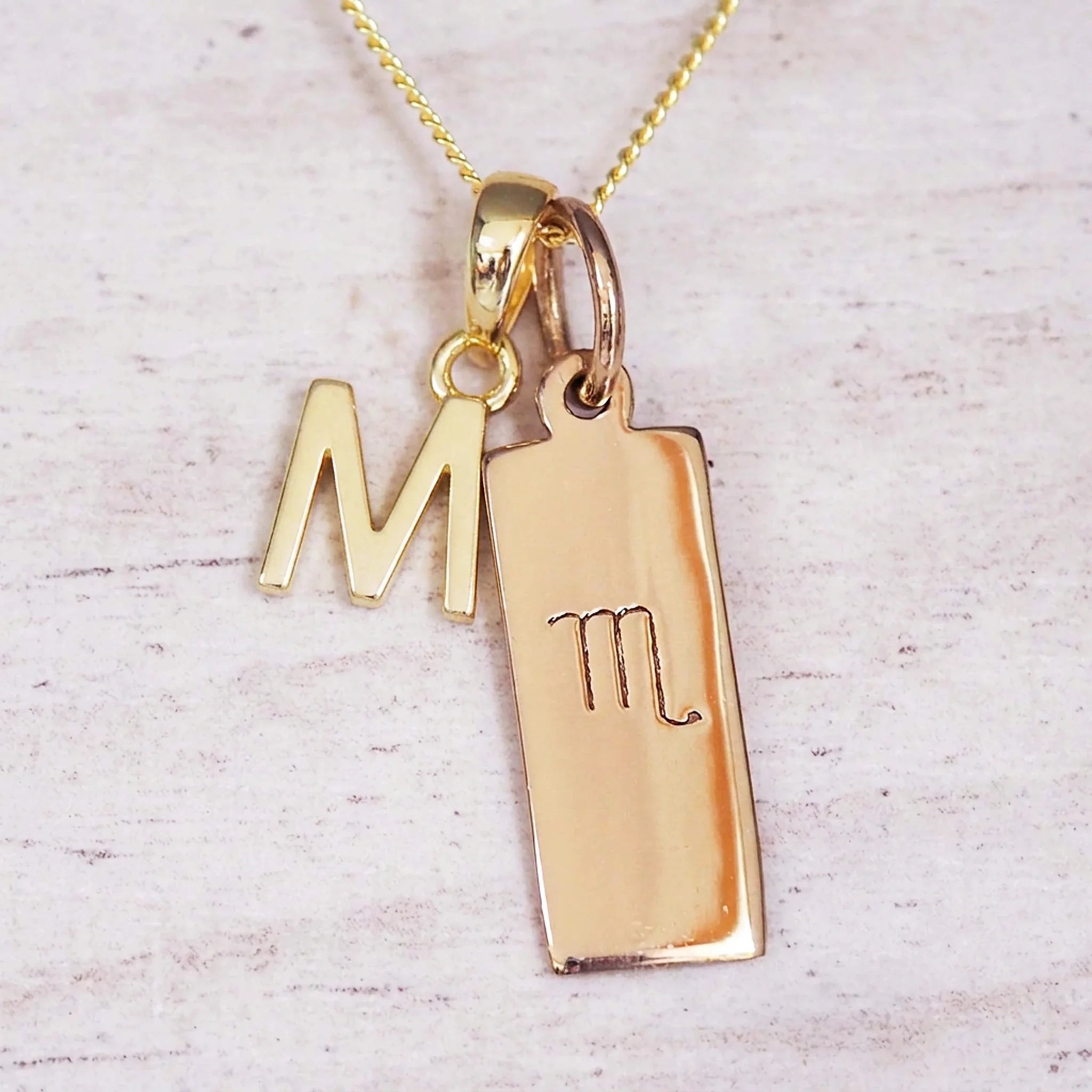 Gold Scorpio initial necklace with a letter M charm and engraved zodiac bar on white wood, close-up