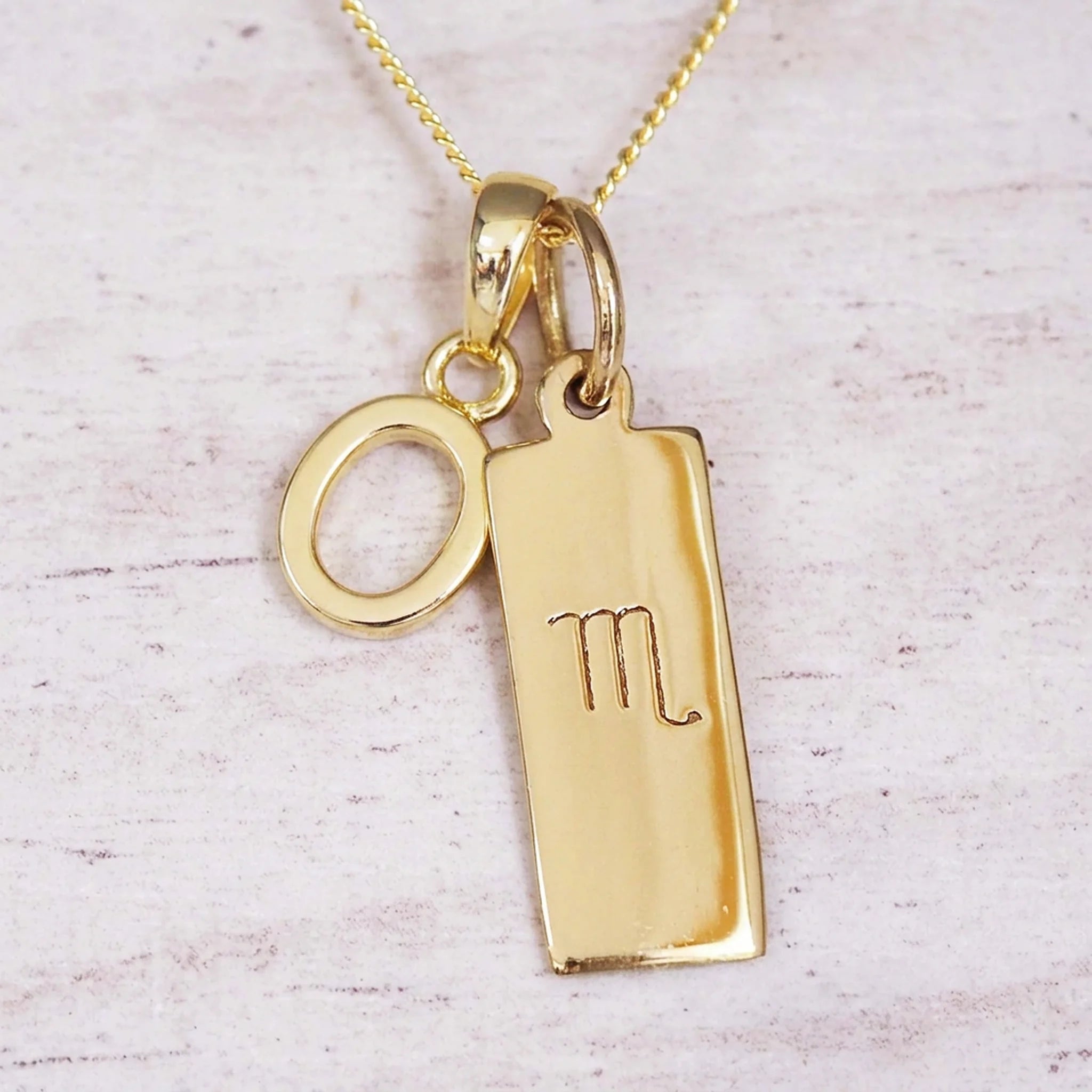 Gold Scorpio initial necklace with a letter O charm and engraved zodiac bar on white wood, close-up
