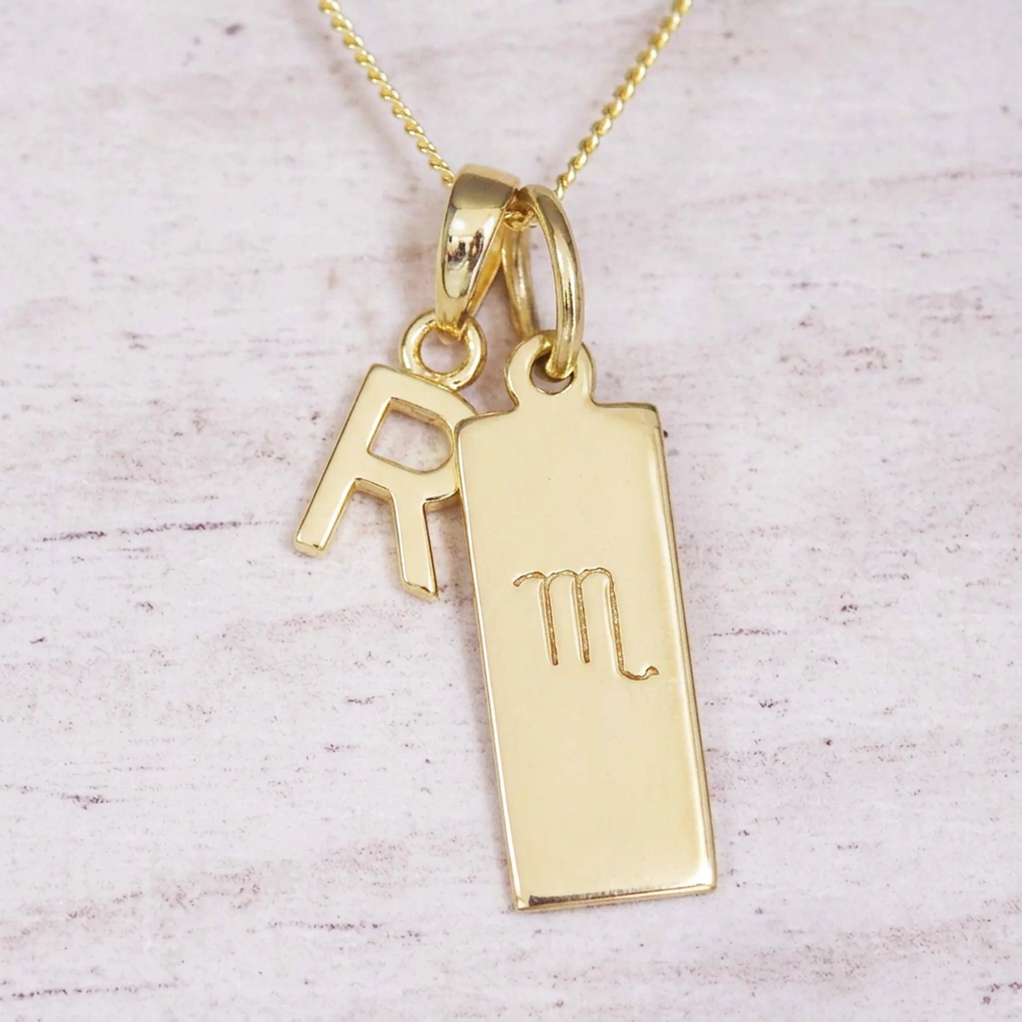 Gold Scorpio initial necklace with a letter R charm and engraved zodiac bar on white wood, close-up
