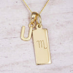 Gold Scorpio initial necklace with a letter U charm and engraved zodiac bar on white wood, close-up