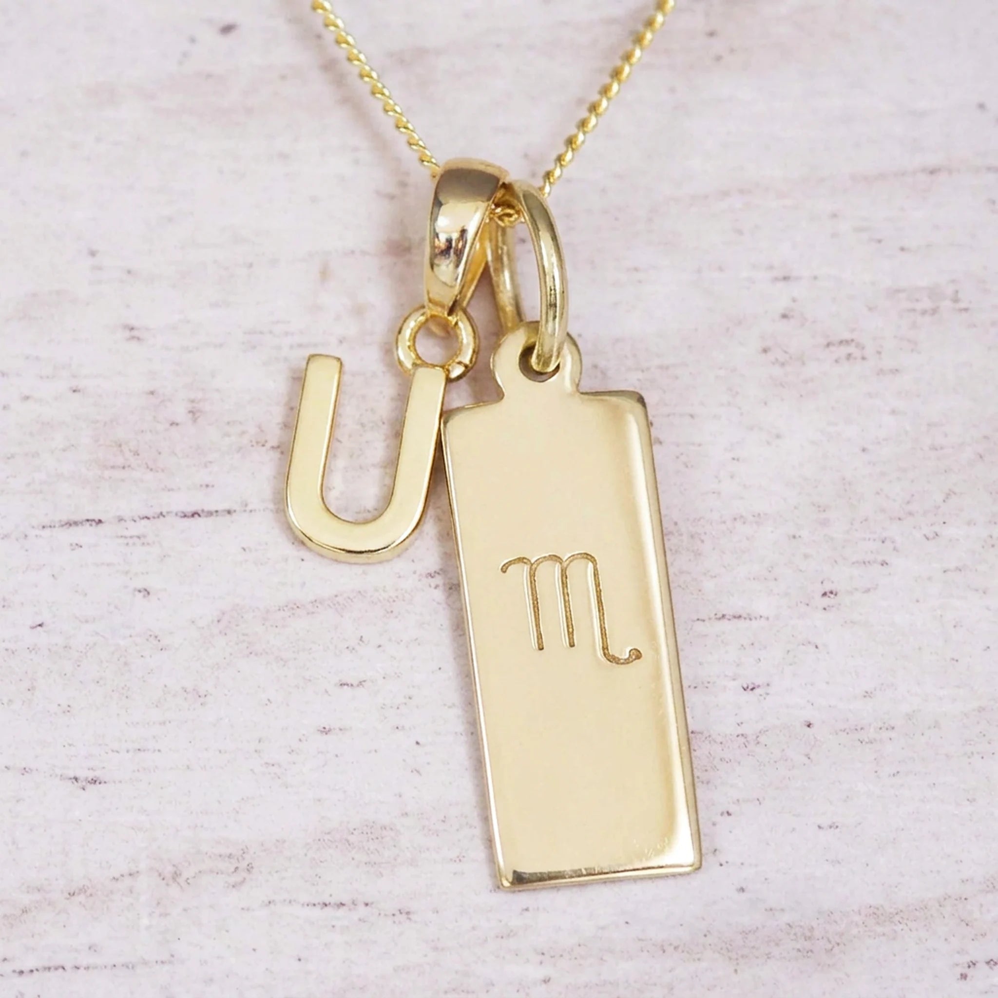 Gold Scorpio initial necklace with a letter U charm and engraved zodiac bar on white wood, close-up