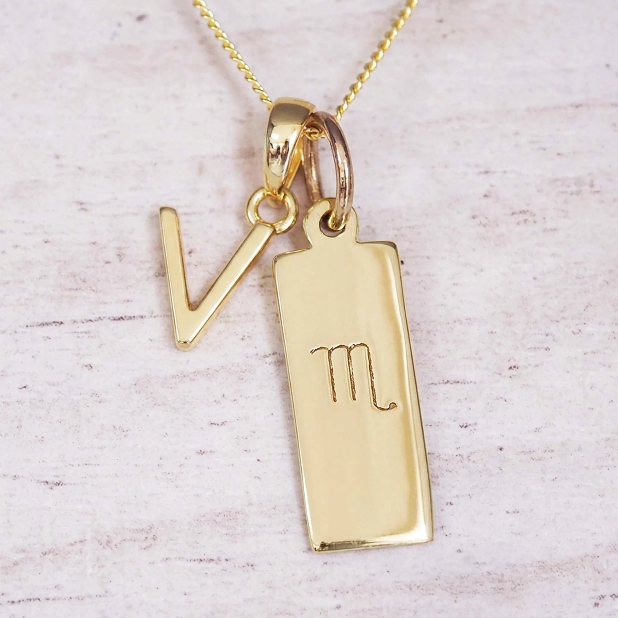 Gold Scorpio initial necklace with a letter V charm and engraved zodiac bar on white wood, close-up