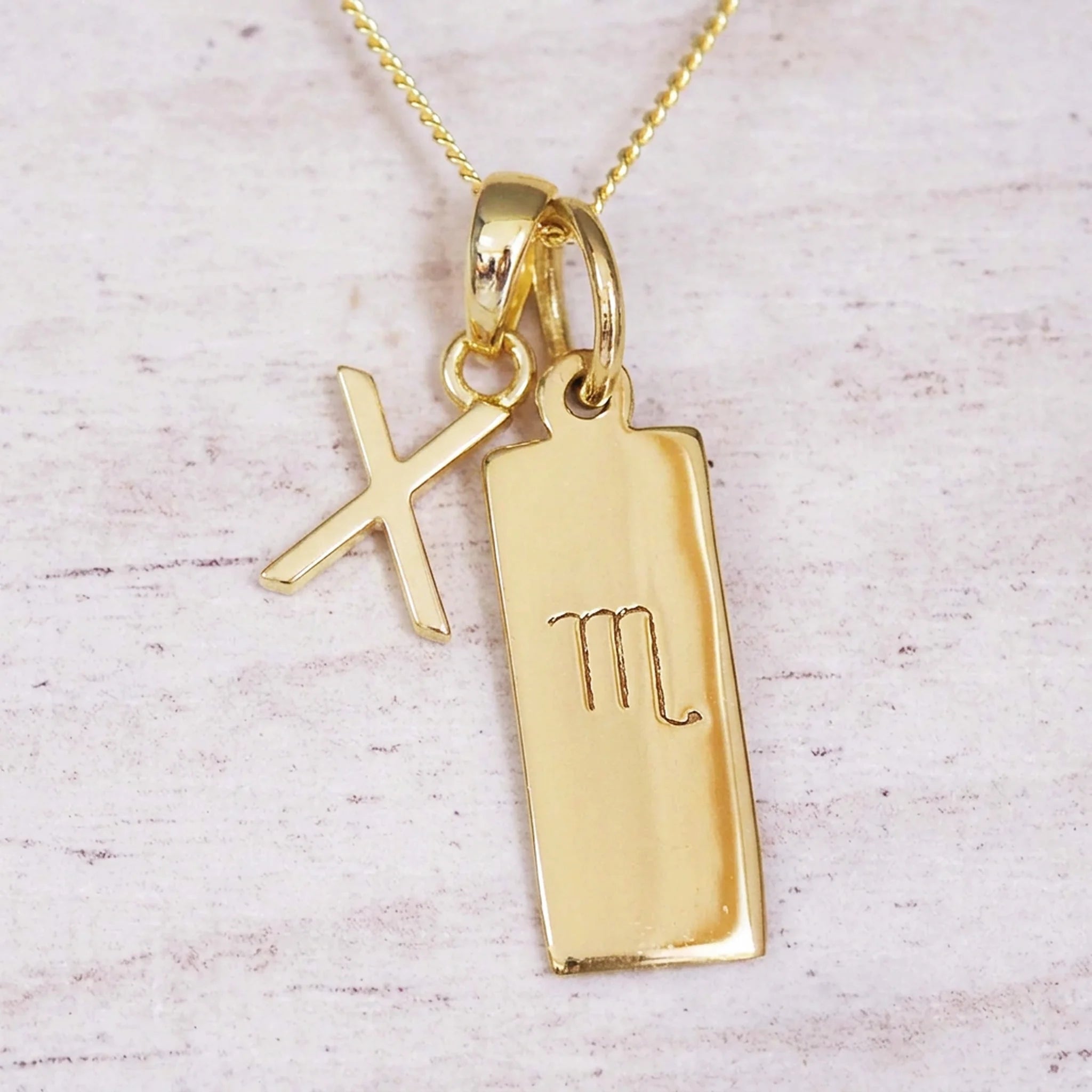 Gold Scorpio initial necklace with a letter X charm and engraved zodiac bar on white wood, close-up