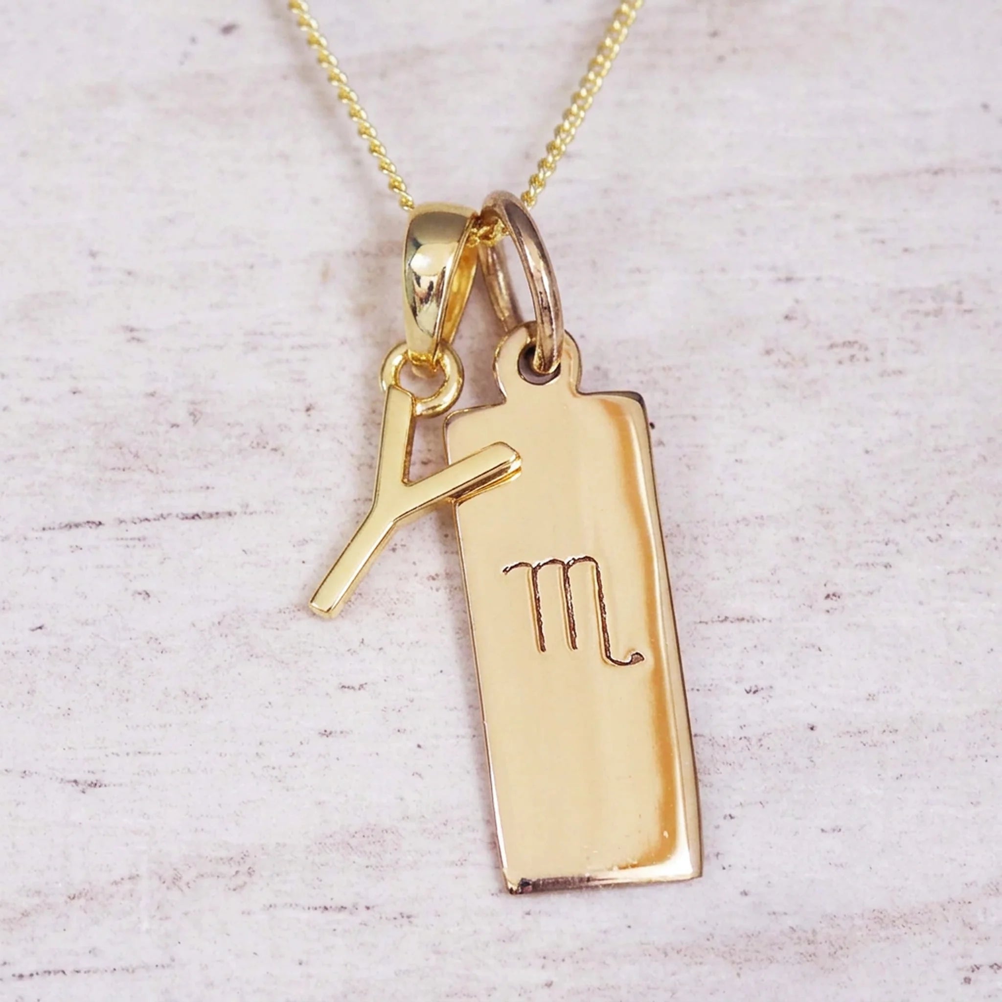 Gold Scorpio initial necklace with a letter Y charm and engraved zodiac bar on white wood, close-up