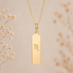 Gold necklace with a rectangular pendant featuring an engraved scorpio zodiac line glyph symbol on a beige background.