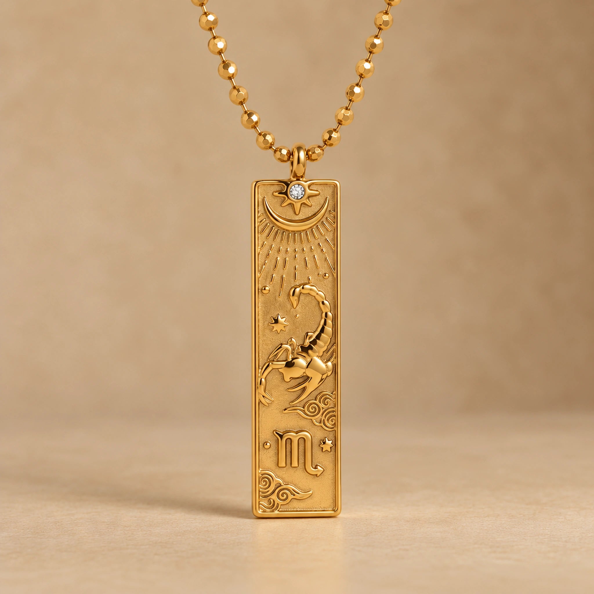 Gold Scorpio zodiac necklace, embossed bar pendant with sign symbol on a faceted ball chain, warm beige background