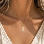 Gold necklace with a rectangular pendant with scorpio glyph symbol on a person wearing a white top.