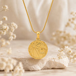 A gold pendant necklace with a circular scorpio zodiac sign design, resting on a textured stone with soft dried flowers in a warm, neutral setting.