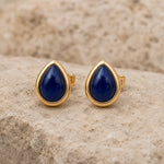 A pair of September Birthstone Natural Sapphire Earrings - gold teardrop stud earrings with deep blue cabochon stones, displayed on a textured beige stone surface.