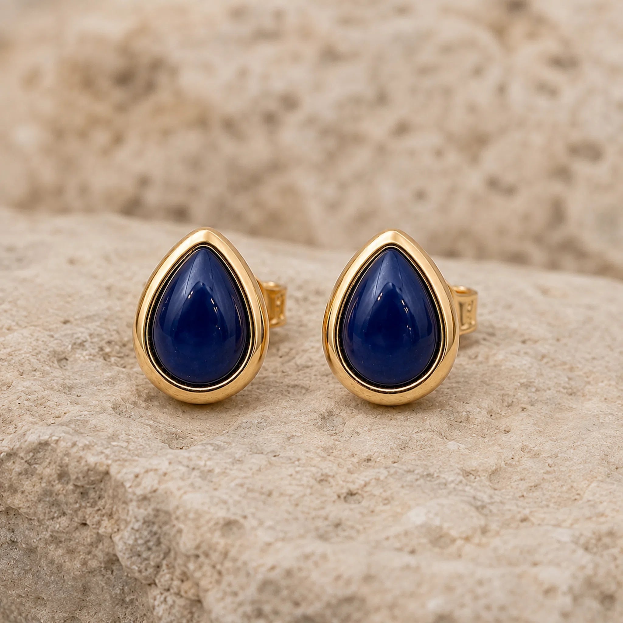 A pair of September Birthstone Natural Sapphire Earrings - gold teardrop stud earrings with deep blue cabochon stones, displayed on a textured beige stone surface.