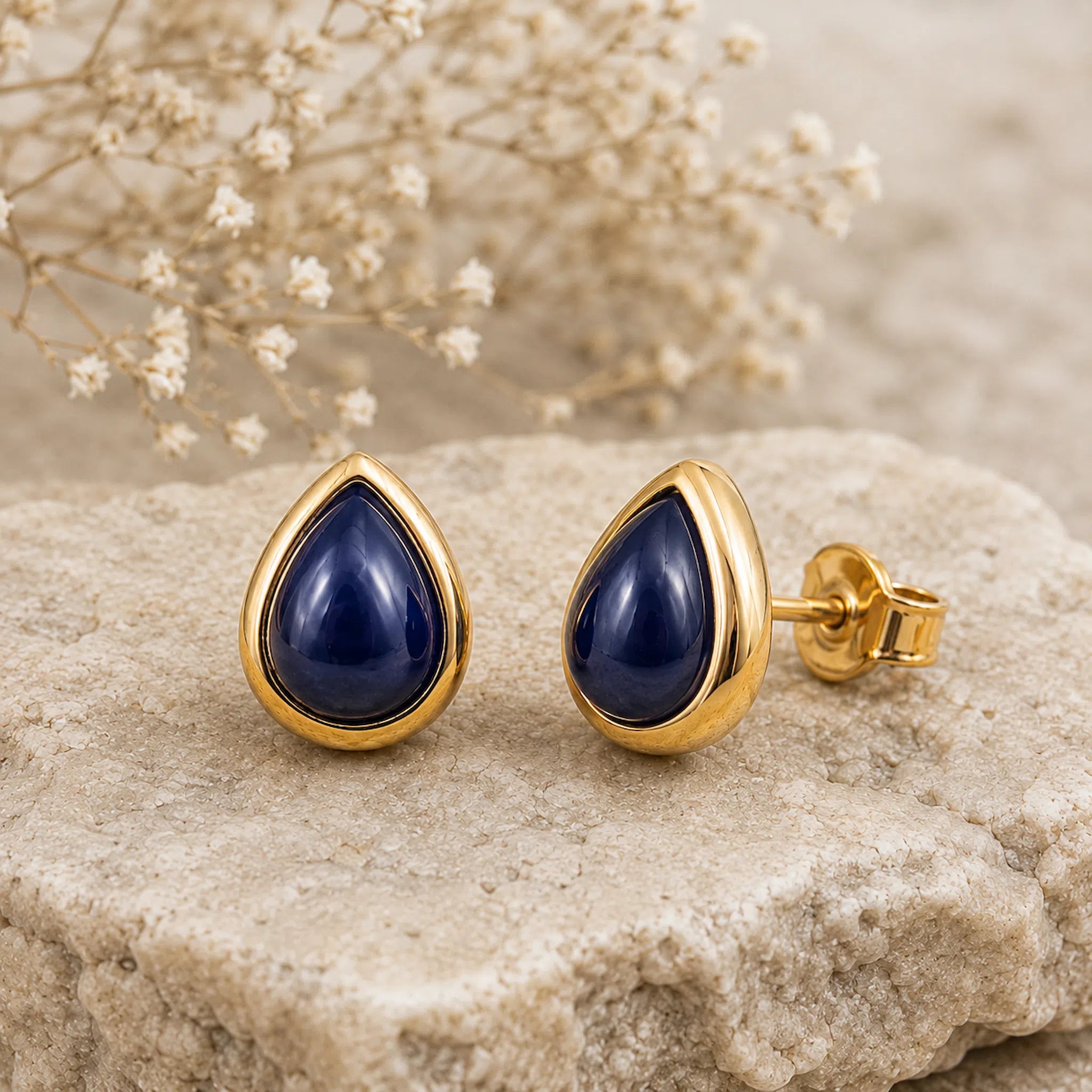 A pair of September birthstone natural Sapphire Earrings - gold teardrop stud earrings with deep navy blue cabochon stones, displayed on a textured light beige stone with dried baby's breath flowers in the background.