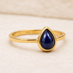september birthstone sapphire ring - A gold ring featuring a teardrop-shaped dark blue lapis lazuli stone in a bezel setting, shown on a textured light beige surface.