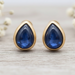 Teardrop-shaped blue sapphire gemstone earrings with gold settings on a light wooden surface.