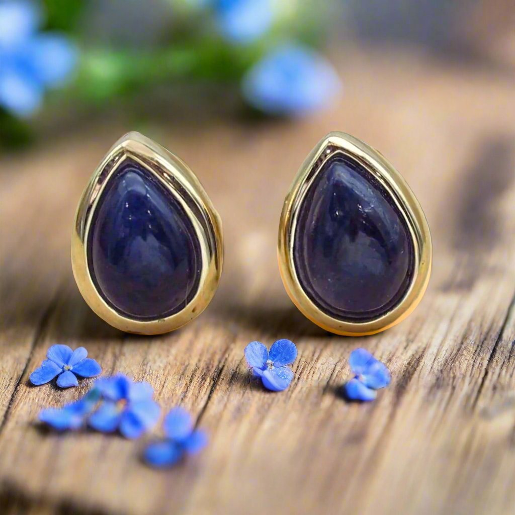 Gold teardrop earrings with sapphire stones on a wooden surface with small blue flowers.