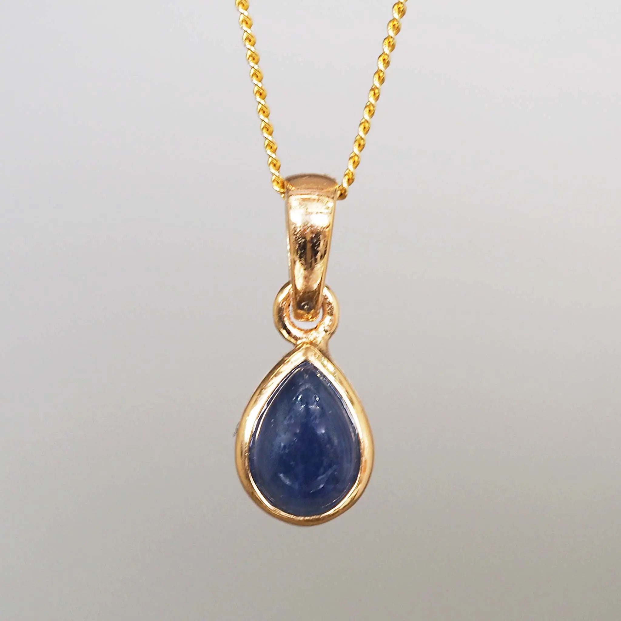 Gold pendant with a tear drop shaped blue sapphire gemstone on a gold chain against a gray background