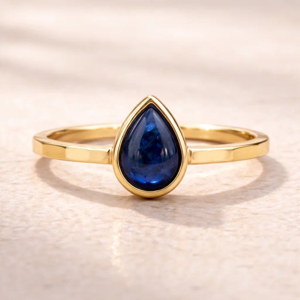 Gold ring with a blue teardrop-shaped natural sapphire gemstone on a beige background