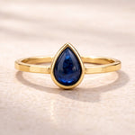 Gold ring with a blue teardrop-shaped natural sapphire gemstone on a beige background